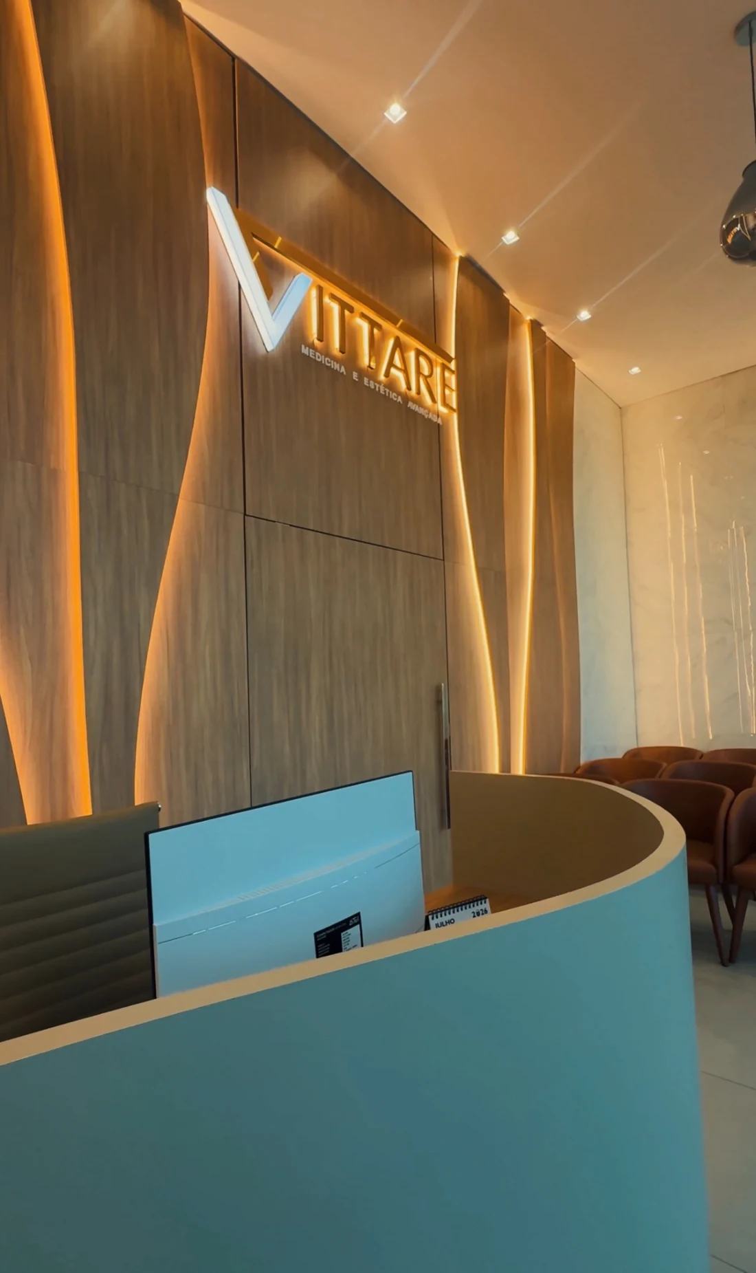 Recepção da Clínica Vittare, com balcão curvo, logo iluminado e revestimento de madeira.
